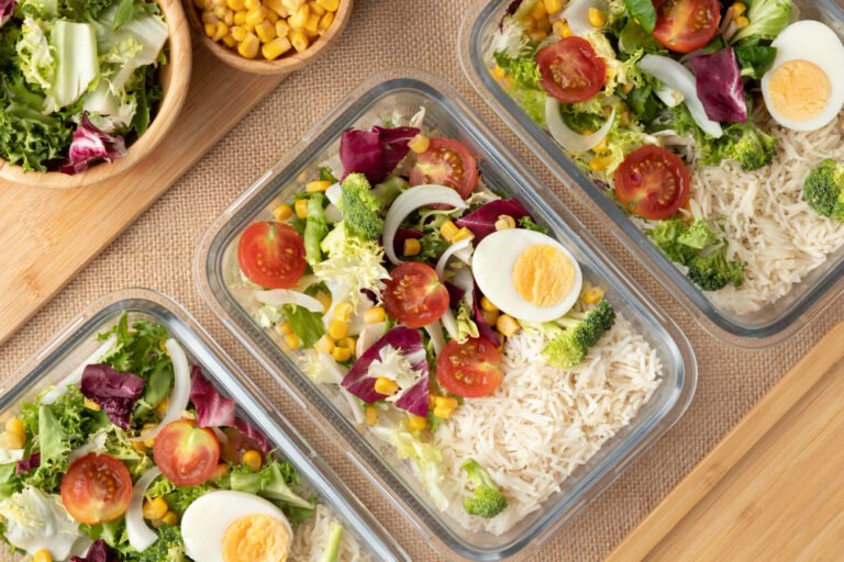 Glass meal prep containers with mixed salad, corn, boiled eggs and cherry tomatoes on wooden board